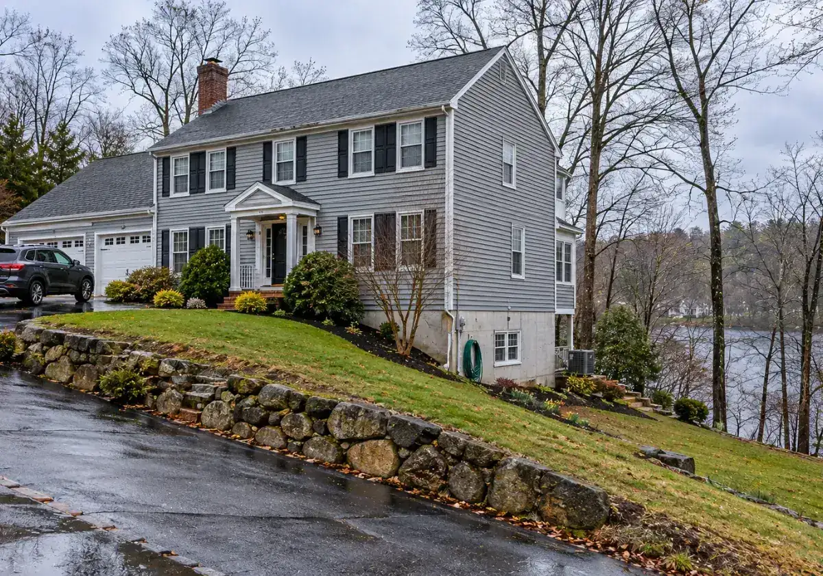 Gray colonial home on a sloped lot with a stone wall overlooking a pond in Cummingsville, Woburn, MA
